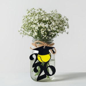 May include: A glass jar filled with water and white flowers. A black graduation cap and yellow tennis ball are attached to the jar with the number 2020. The jar is tied with twine.