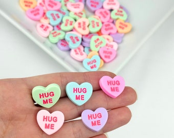 May include: Five pastel-colored heart-shaped candies with the text "Hug Me" in pink lettering. The candies are in a hand.