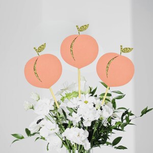 Op de afbeelding: Drie perzikvormige papieren decoraties met gouden glitteraccenten, bevestigd aan houten spiesjes, zijn gerangschikt in een boeket witte bloemen.