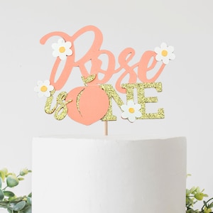 May include: Peach-themed cake topper with the words "Rose is One" in pink and gold glitter. The topper features a peach, flowers, and glitter accents.