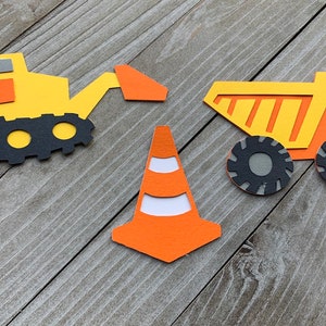 May include: Three construction-themed paper cutouts: a yellow excavator with a black track, a yellow dump truck with orange stripes, and an orange traffic cone with white stripes.