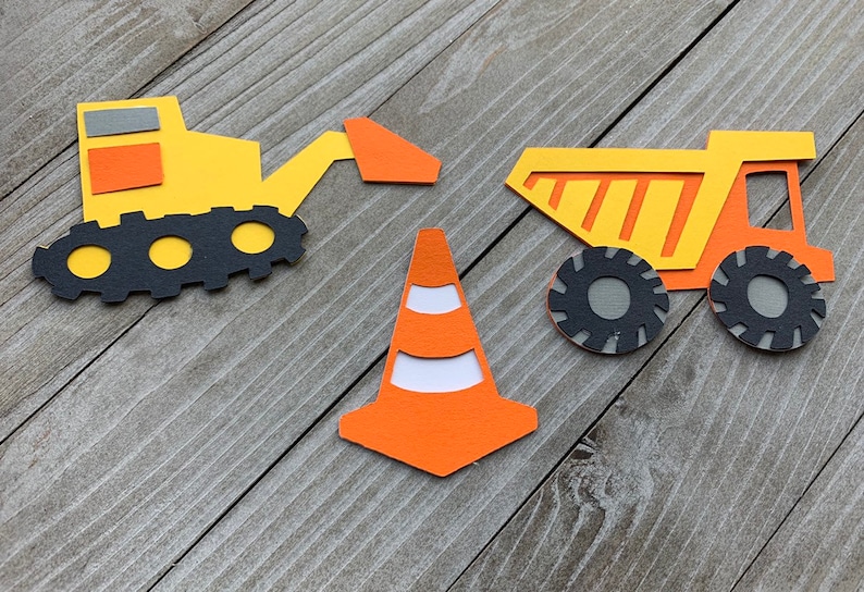 May include: Three construction-themed paper cutouts: a yellow excavator with a black track, a yellow dump truck with orange stripes, and an orange traffic cone with white stripes.
