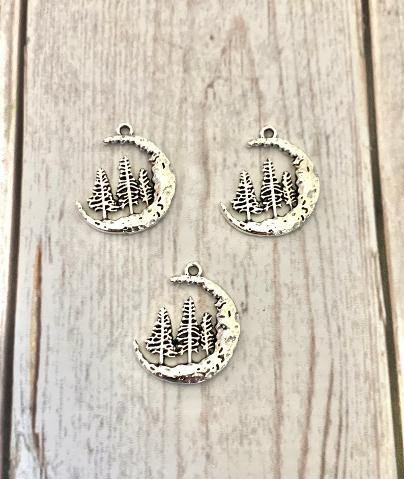 May include: Three silver charm pendants shaped like a crescent moon with three pine trees inside the moon. The moon has a textured surface.