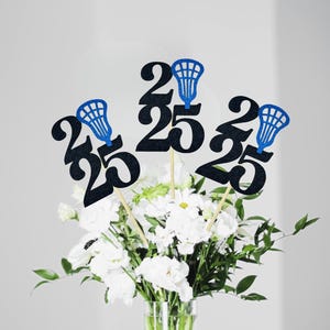 May include: Black glittery number 25 centerpieces with blue lacrosse stick accents on wooden sticks. The centerpieces are arranged in a vase of white flowers and greenery, perfect for a graduation celebration.