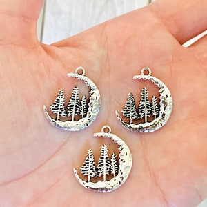 May include: Three silver charm pendants featuring a crescent moon with three stylised pine trees inside. The moon has a textured surface.