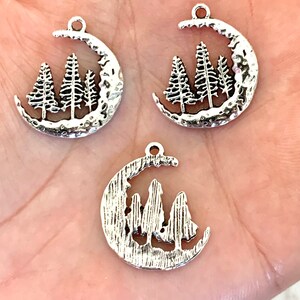 May include: Three silver charm pendants featuring a crescent moon with a silhouette of three pine trees inside. The moon has a textured surface.