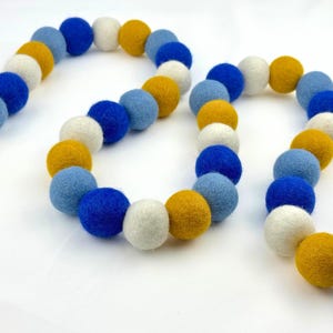 May include: A colorful felt ball garland in shades of blue, white, and yellow. The garland is made of small, round felt balls strung together. The garland is a decorative item, suitable for various occasions.