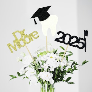 Puede incluir: Centro de mesa de graduación con un gorro de graduación negro, un diente blanco y un 2025 negro con un gorro de graduación. El centro de mesa está decorado con purpurina dorada y se coloca en un jarrón con flores blancas.