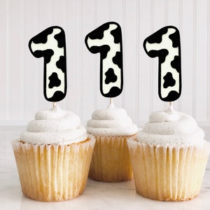 Pode incluir: Três cupcakes com cobertura branca e toppers de número um com estampa de vaca em preto e branco.