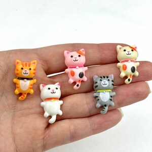 May include: Five small plastic figurines of cats in different colors and patterns. The cats are all in a standing position with their paws raised. The cats are orange, pink, white, gray, and cream.