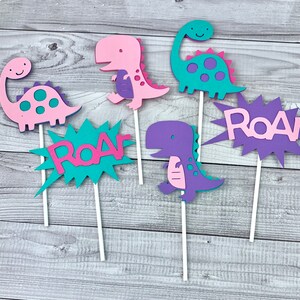 K&ouml;nnte beinhalten: Sechs bunte Dinosaurier- und "Roar"-Tortenaufleger. Die Aufleger sind aus Schaumstoff und in den Farben Pink, Lila, T&uuml;rkis und Gr&uuml;n gehalten.