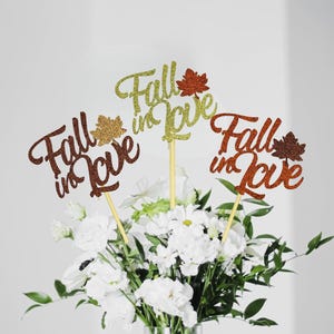 May include: Three gold glitter "Fall in Love" signs with a leaf design, each on a wooden stick, in a bouquet of white flowers.