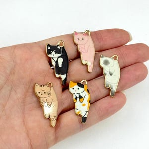Könnte beinhalten: Fünf goldfarbene Metall-Anhänger in Katzenform. Die Anhänger sind mit verschiedenen Katzendesigns bemalt, darunter schwarz-weiß, rosa, orange-weiß und weiß. Die Anhänger hängen an einer Hand.