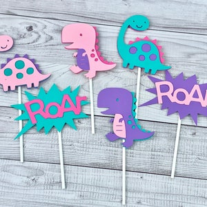 K&ouml;nnte beinhalten: Sechs bunte Dinosaurier-Kuchentopper, darunter drei Dinosaurier und drei "Roar"-Schilder. Die Dinosaurier sind rosa, lila und t&uuml;rkis, und die Schilder sind t&uuml;rkis und lila. Die Topper sind perfekt f&uuml;r eine Dinosaurier-Party.