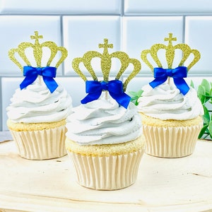 Könnte beinhalten: Drei Cupcakes, die mit goldenen Glitzerkronen und blauen Schleifen verziert sind. Die Cupcakes stehen auf einer Holzoberfläche.