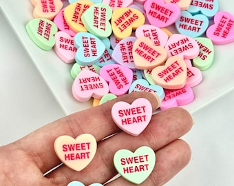 May include: Assortment of colorful heart-shaped candies with the words "Sweet Heart" printed on them. The candies are arranged in a pile on a white surface.