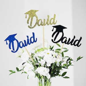 May include: Graduation-themed floral arrangement with three glittery toppers spelling "David" and a graduation cap. The toppers are gold, blue, and black, and are set in a vase of white flowers and greenery. A celebratory decoration.
