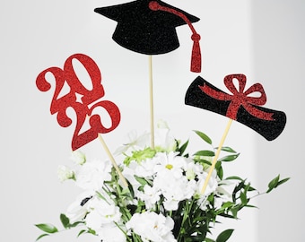 Puede incluir: Un gorro de graduación, un diploma y los números "2023" de purpurina roja en palos, dispuestos en un ramo de flores blancas.