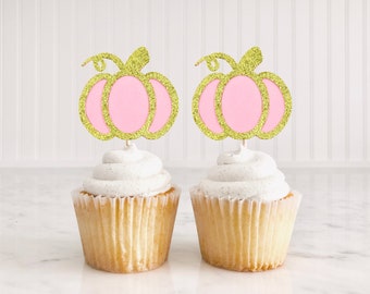 May include: Two cupcakes topped with white frosting and decorated with pink and gold glitter pumpkin cupcake toppers.