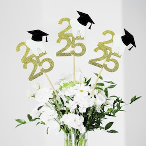 Puede incluir: Tres centros de mesa de graduación de purpurina dorada con el número 2025. Cada centro de mesa presenta un diente blanco con un gorro de graduación negro en la parte superior. Los centros de mesa están dispuestos en un ramo de flores blancas.