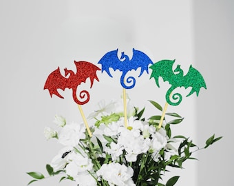 May include: Three glitter dragon cupcake toppers in red, blue, and green. The toppers are on wooden sticks and are inserted into a bouquet of white flowers.
