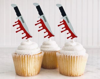 Può includere: Tre cupcake bianchi con decorazioni a forma di coltello rosso e grigio. I coltelli hanno sangue rosso che cola. I cupcake sono su una superficie bianca.