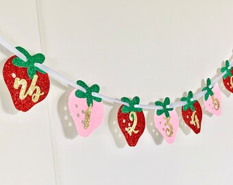 May include: A pink and red glitter strawberry banner with gold numbers 1 through 6 and the word "one" in gold letters. The banner is strung on white string.