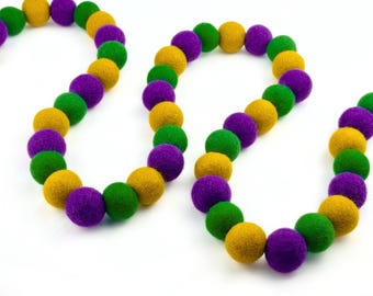 May include: A colorful garland featuring felt balls in green, gold, and purple. The decorative string of balls is arranged in a curved pattern against a white background. The garland is suitable for festive occasions.