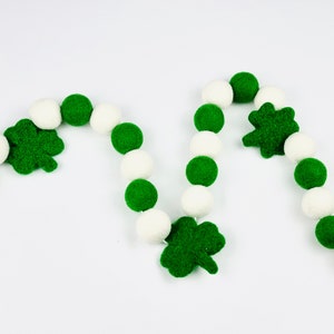 Peut inclure: Une guirlande festive composée de boules de feutre blanches et vertes alternées, parsemée de motifs de trèfles et d'étoiles verts. Conçue pour les décorations de la Saint-Patrick, elle se détache sur un fond blanc.