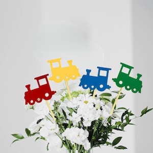 Puede incluir: Un ramo de flores blancas y follaje verde con cuatro decoraciones coloridas en forma de tren sobre palitos. Los trenes son rojos, amarillos, azules y verdes. Decoraciones para una fiesta o evento.