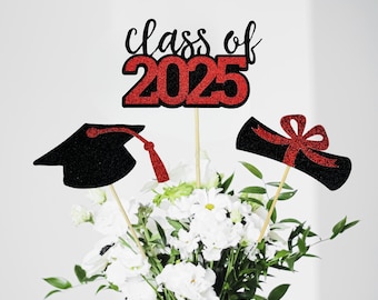 Puede incluir: Tres palos de centro de mesa para una fiesta de graduación. Un palo tiene un gorro de graduación negro, otro tiene un letrero de "Clase de 2025" de brillo rojo y el último tiene un rollo de graduación negro con un lazo rojo.
