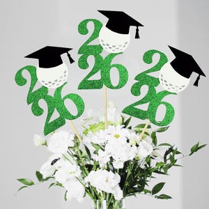 Puede incluir: Centros de mesa de graduación con purpurina verde con el número "26" y un gorro de graduación sobre una pelota de golf. Los centros de mesa están colocados en un jarrón con flores blancas y vegetación.
