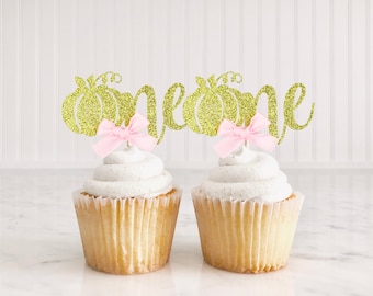 Peut inclure: Deux cupcakes avec un glaçage blanc surmontés de décorations de cupcake en or pailleté "un" avec un nœud rose. Les décorations présentent un motif de citrouille.