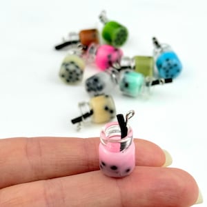Op de afbeelding: Een verzameling miniatuur bubble tea-charms in verschillende kleuren, waaronder roze, blauw, groen en bruin. Elke charm heeft een doorzichtige plastic fles gevuld met een gekleurde vloeistof en zwarte boba-parels, en een klein zwart rietje.