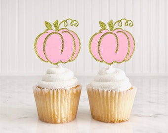 Peut inclure: Deux cupcakes avec un glaçage blanc surmontés de décorations de cupcakes en forme de citrouille rose pailletée. Les citrouilles ont des tiges et des feuilles pailletées dorées.