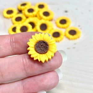 May include: A close-up of a yellow sunflower with a brown centre. The sunflower is made of plastic and is about 1 inch in diameter. There are other sunflowers in the background.