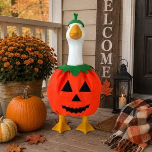 May include: A white goose figurine wearing a bright orange pumpkin costume with a black jack-o'-lantern face and a green stem hat. The goose has a yellow beak and yellow webbed feet, standing upright as a decorative piece for seasonal display.