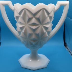 May include: A white milk glass vase featuring a geometric diamond pattern across the body, a scalloped rim, and two symmetrical handles extending from the sides. The vessel stands on a pedestal base with an octagonal shape and smooth finish.