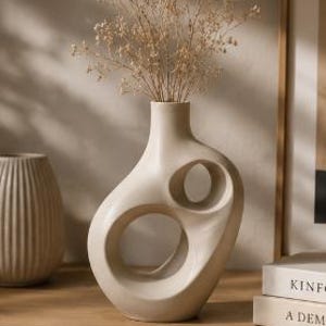 Peut inclure: Un vase en céramique beige avec un design organique unique, comprenant deux grandes découpes circulaires. Le vase contient des tiges décoratives séchées, de couleur brun clair. Un vase plus petit et nervuré est partiellement visible à gauche. Le vase est sur une surface en bois.