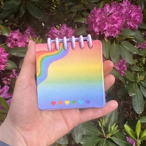 May include: A small square notepad with a spiral binding at the top. The cover features a rainbow gradient background with a wavy rainbow stripe in the upper left corner and a row of six small, colorful heart icons printed along the bottom edge.