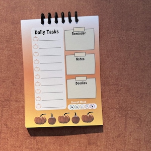 May include: A spiral-bound daily planner notepad with an orange and yellow gradient background. The page features sections for Daily Tasks with pumpkin icons, Reminder, Notes, Doodles, and an Overall Mood tracker with five emoji faces.