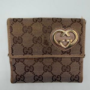 May include: A brown and beige bi-fold wallet featuring the iconic interlocking GG monogram pattern across the fabric surface. The front flap is adorned with a prominent gold-toned metal emblem shaped like a heart with the Gucci logo inside.