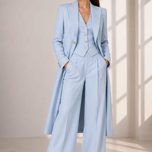Puede incluir: Un traje de tres piezas azul claro que incluye un blazer largo, un chaleco abotonado con botones dorados y pantalones de pierna ancha. El nombre de la marca TUXORA aparece en la esquina superior izquierda. Se combina con zapatos blancos.