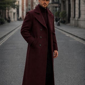 Puede incluir: Un abrigo largo de lana color burdeos con doble botonadura, solapas anchas y botones oscuros, sobre un suéter de cuello alto oscuro. El conjunto incluye una gorra plana de tweed marrón y zapatos de vestir de cuero negro. Se lee la palabra TUXORA.