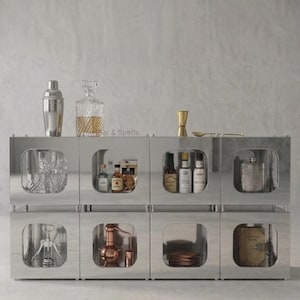 Può includere: Un'unità di stoccaggio modulare in metallo argentato con otto scomparti quadrati dagli angoli arrotondati. L'unità contiene vari accessori da bar, tra cui uno shaker, un decanter, un misurino, un cucchiaio, bicchieri, bottiglie, un cavatappi e un libro.