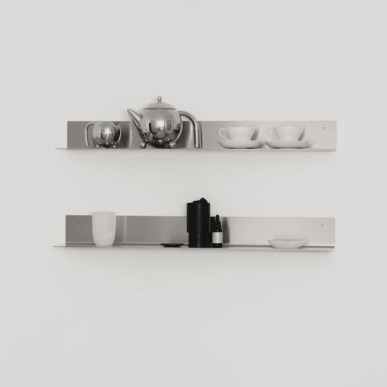 May include: Two horizontal brushed metal wall shelves. The top shelf holds a silver teapot, a small silver creamer, and two white teacups on saucers. The bottom shelf holds a white cup, a black cylindrical container, a small bottle, and a white bowl.