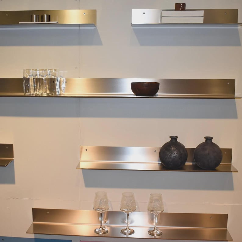 May include: A series of brushed metal wall-mounted shelves arranged in a staggered layout. The shelves hold various items including clear glass tumblers, a dark brown bowl, two round dark gray vases, and three stemmed wine glasses.