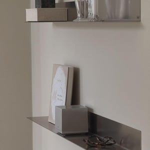 May include: Two brushed metal wall shelves mounted one above the other. The top shelf holds a dark stone cube, a beige fabric box, and a clear glass vase. The bottom shelf holds a beige book, a silver square speaker, and tortoise shell glasses.