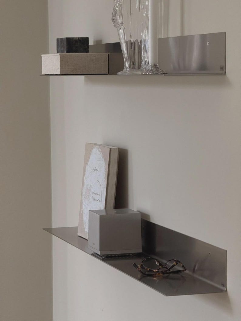 May include: Two brushed metal wall shelves mounted one above the other. The top shelf holds a dark stone cube, a beige fabric box, and a clear glass vase. The bottom shelf holds a beige book, a silver square speaker, and tortoise shell glasses.