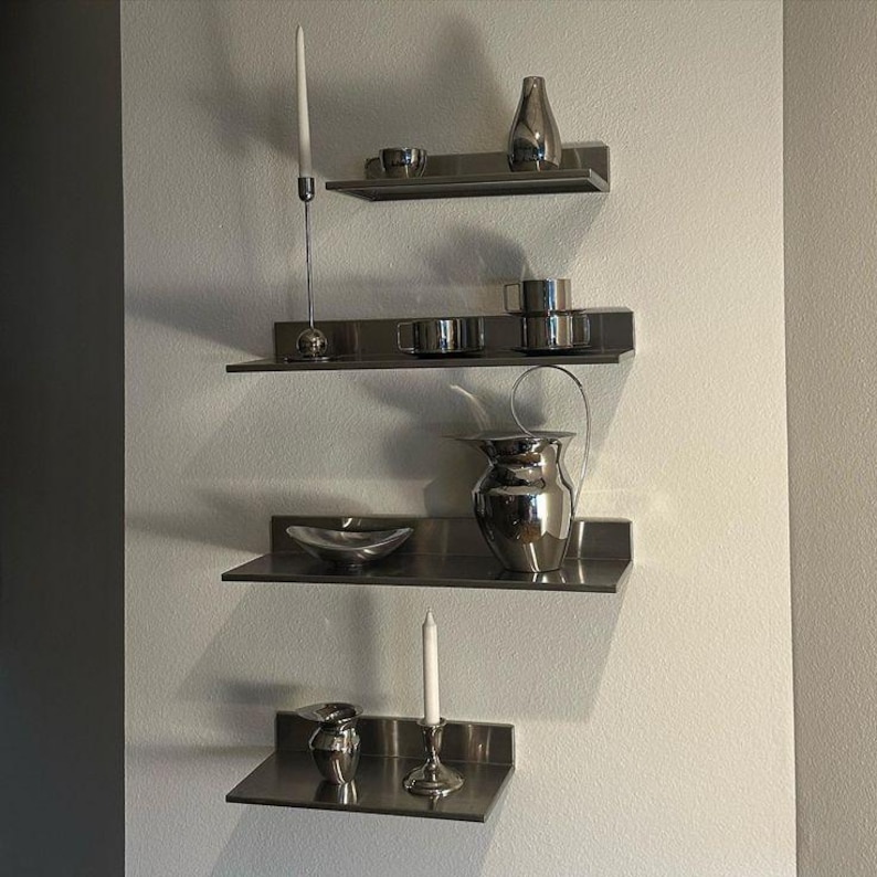 May include: Four horizontal stainless steel wall shelves arranged vertically. The shelves hold various silver-toned items including two tall white taper candles in metal holders, a small vase, a bowl, a pitcher with a handle, and several cups.